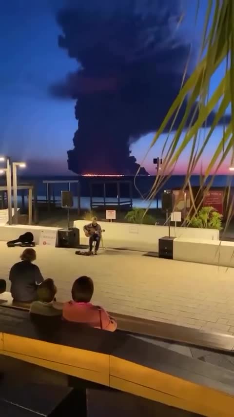 A Sotchi un musicien de rue continue de jouer de la musique sur le front de mer pendant qu'un pétrolier brûle au large en arrière-plan. (métaphore)